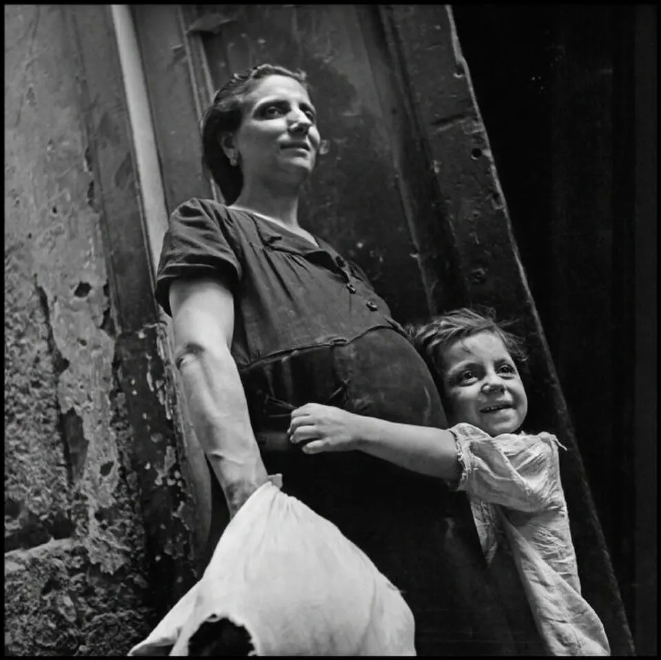 Fotografia storica in bianco e nero che ritrae una donna e un bambino sorridente che la stringe in un abbraccio, ambientata all'aperto nei pressi di un edificio d'epoca.