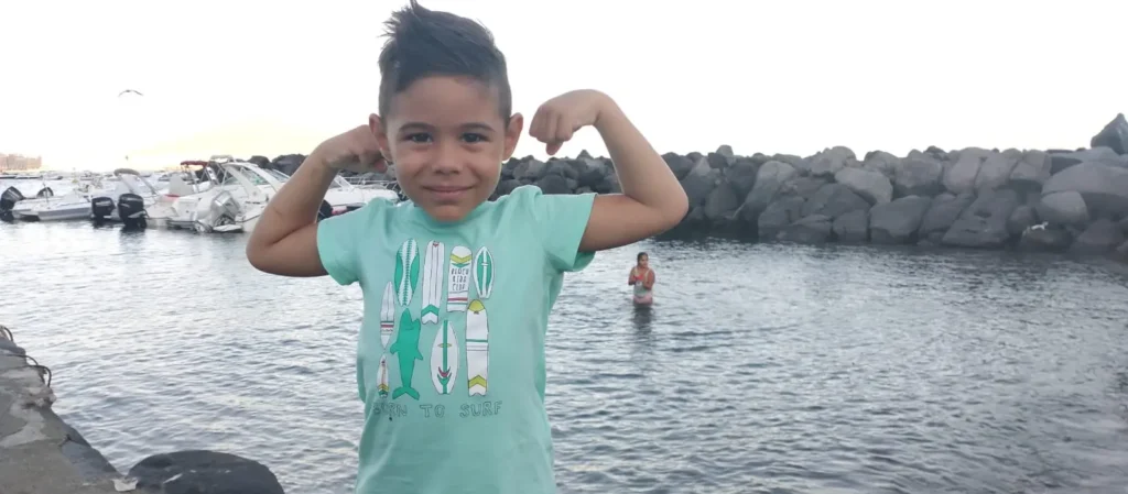 Il piccolo Nicolò sorride e flette i bicipiti in una posa da uomo forte, indossando una maglietta verde menta con tavole da surf, a Mergellina con barche e scogliera sullo sfondo.
