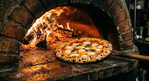 La pizza a Napoli, con cornicione alto e maculato, appena estratta con una pala in legno da un forno a legna in pietra con fiamme vive.